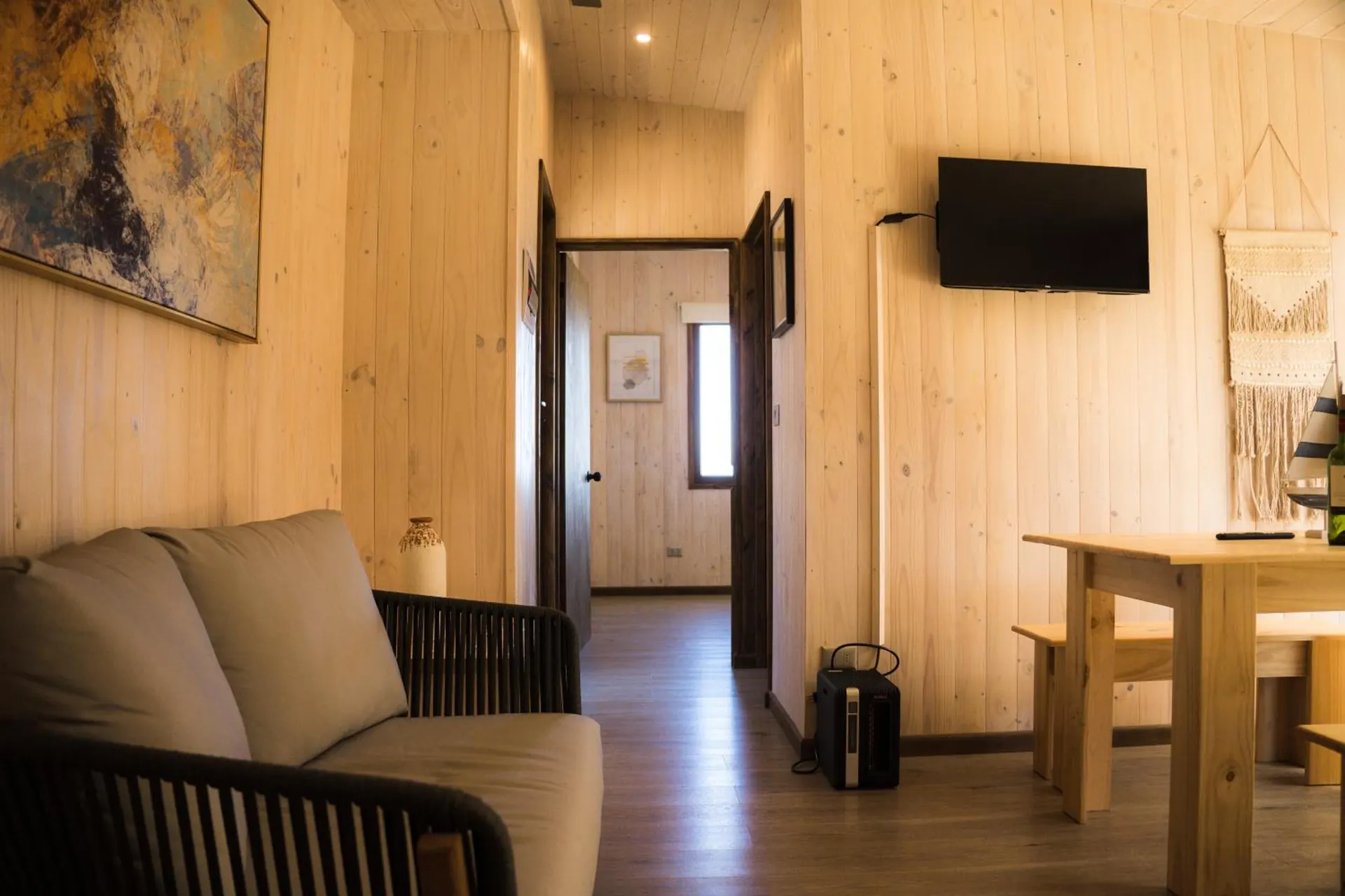 Sala de una cabaña con sofá, televisor en la pared y el comedor al fondo, todo de madera clara.