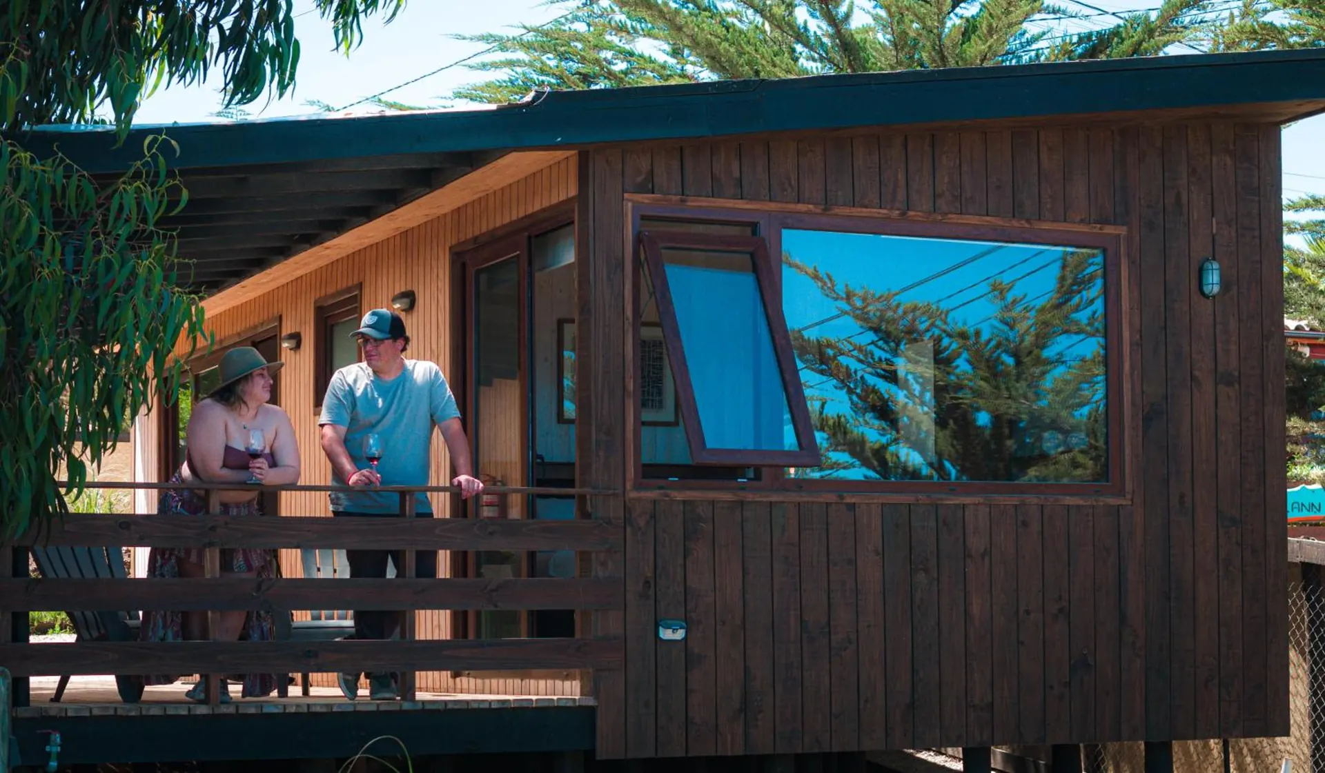Two people chat on the terrace of a dark wood cabin among trees.