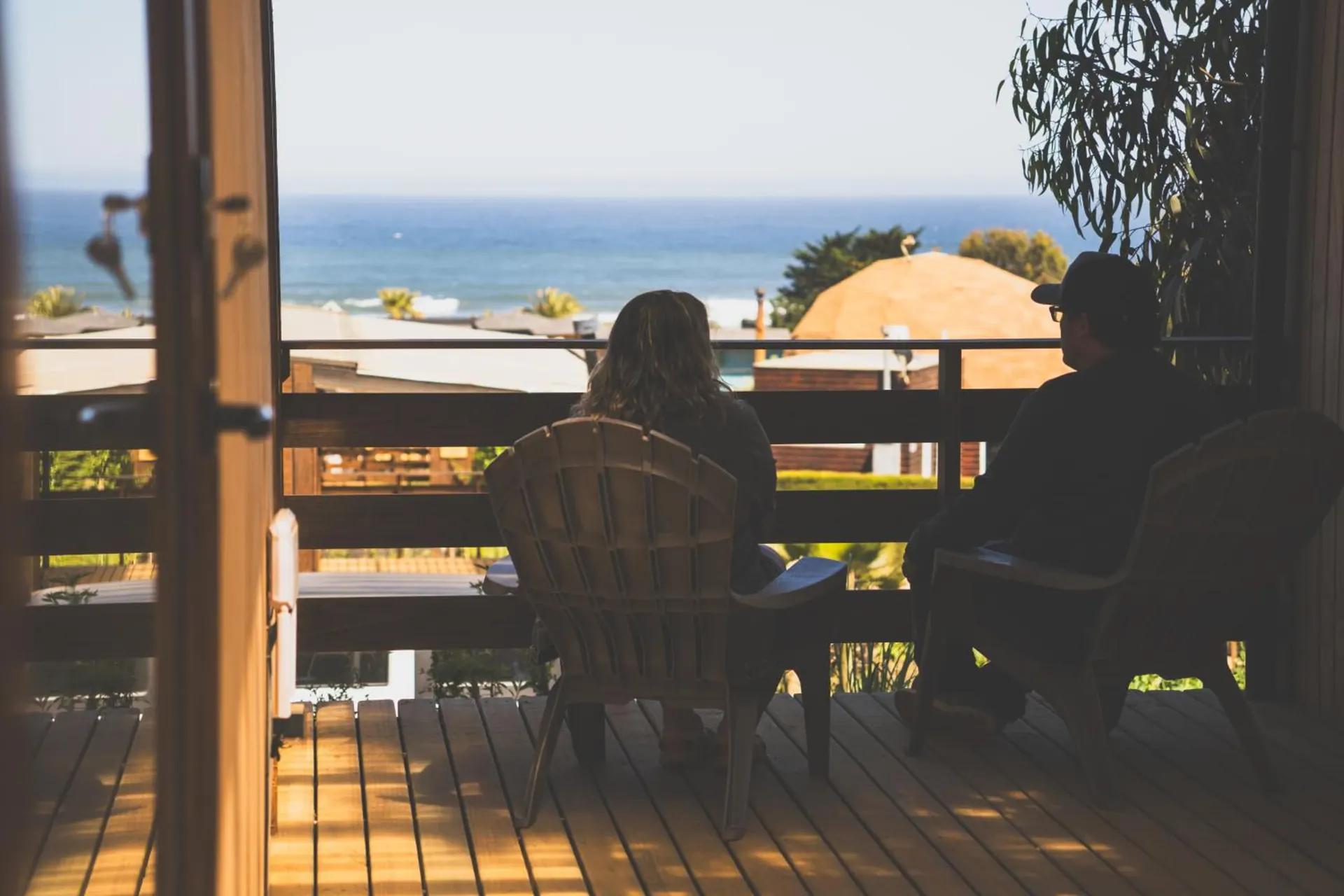 Two people seated on wooden chairs on the cabin terrace, looking at the sea.