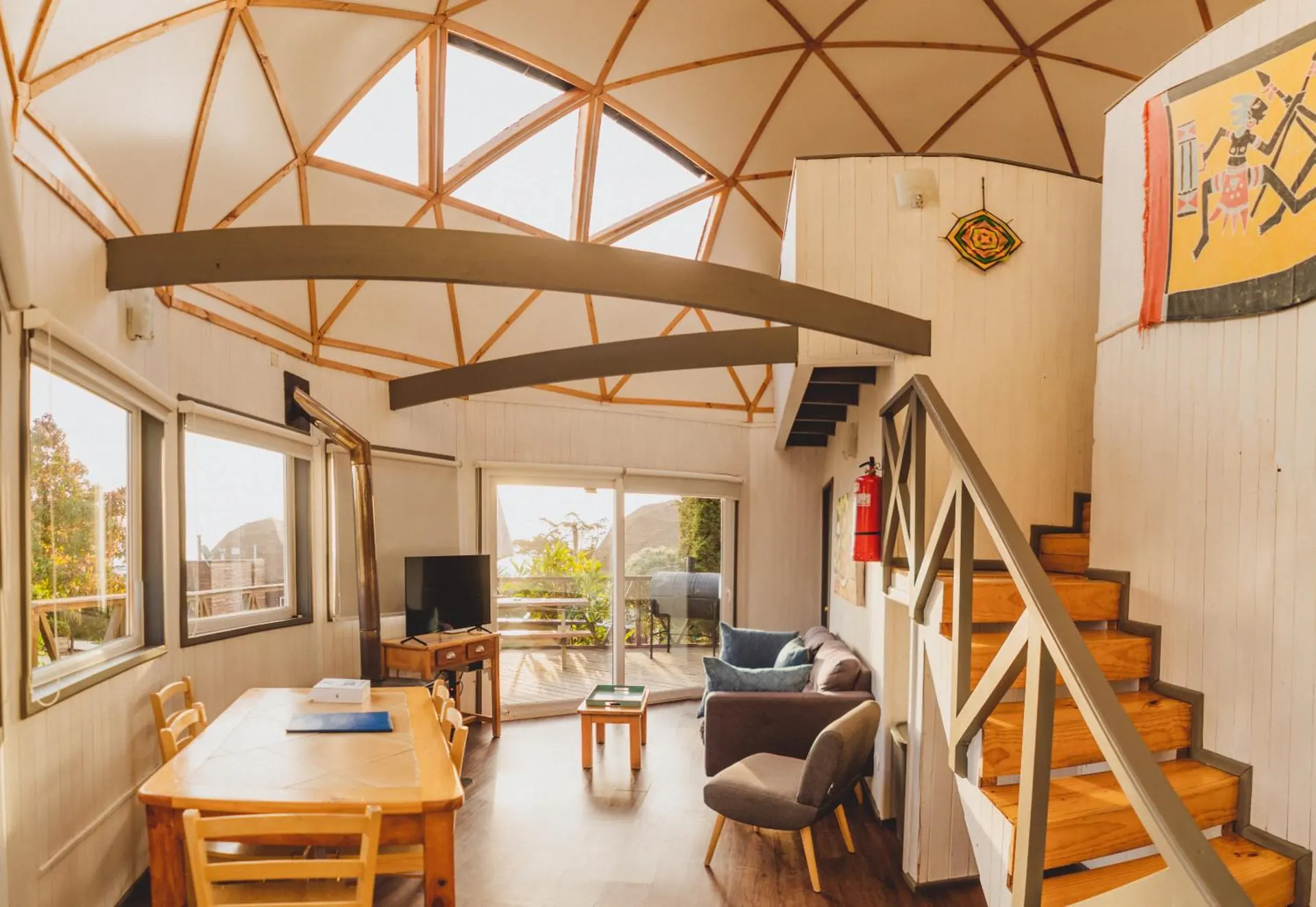 Interior de un domo: techo de vigas en triángulos, escalera de madera, comedor y ventanales.