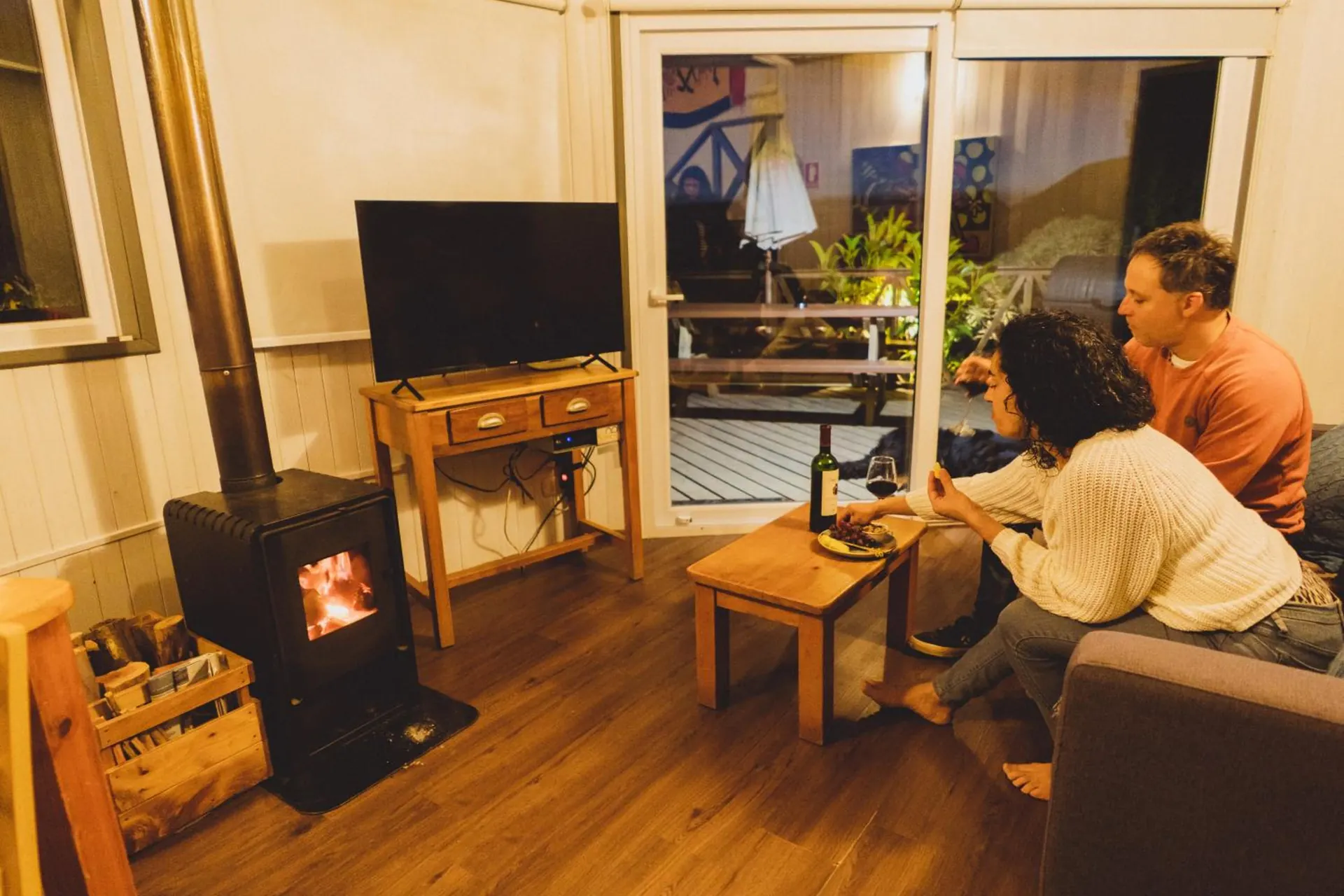 Living room of a dome with a wood stove, a television and a couple seated with a glass of wine.