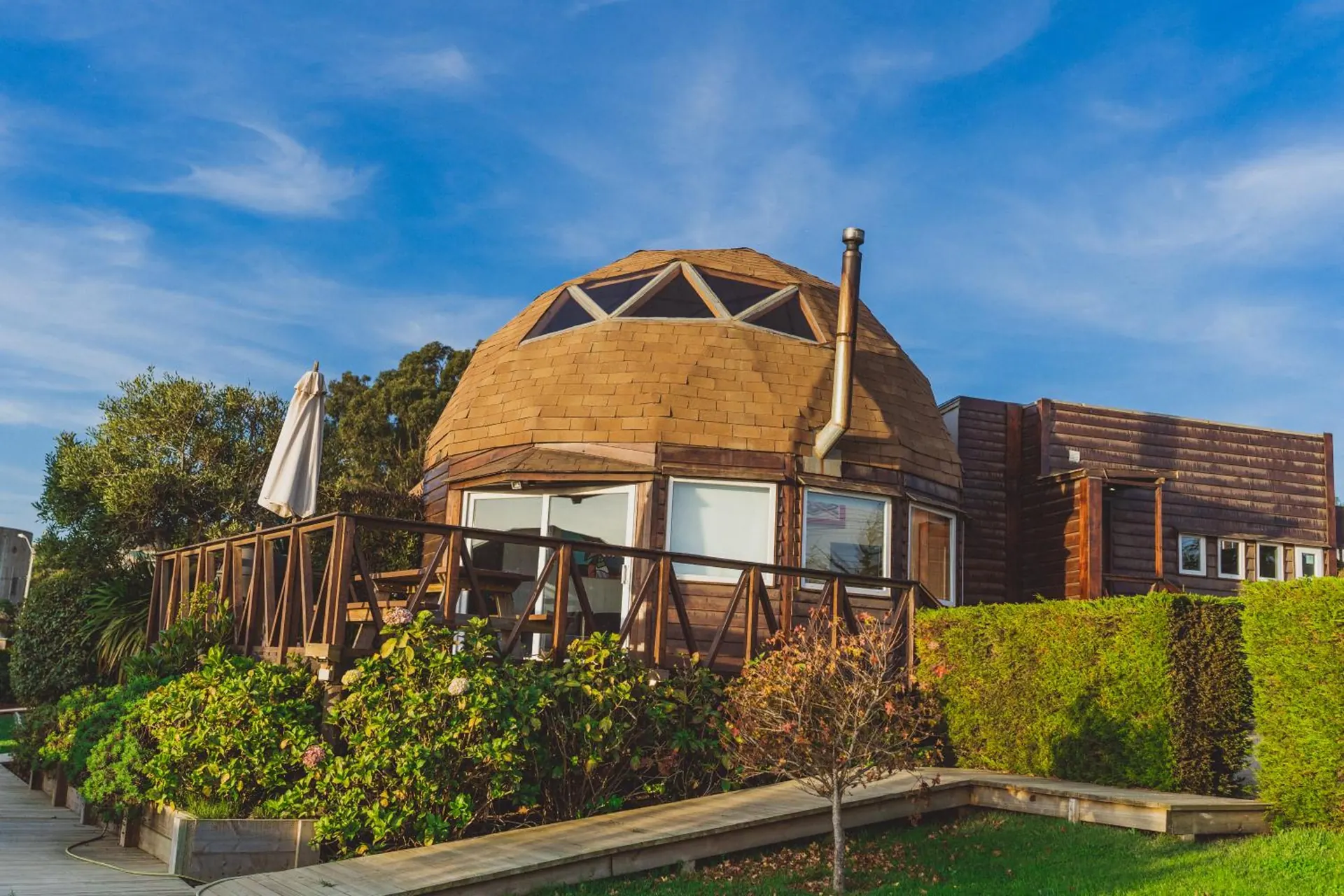 A wooden dome with a terrace and a parasol, among gardens and trees, under a blue sky.
