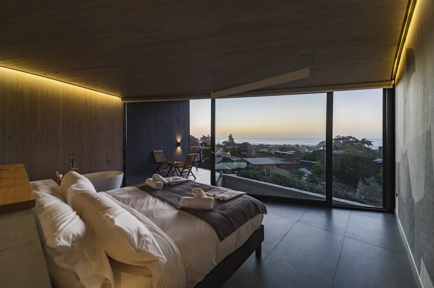 Suíte com cama de casal diante de uma janela do piso ao teto, com o mar e o céu do anoitecer.