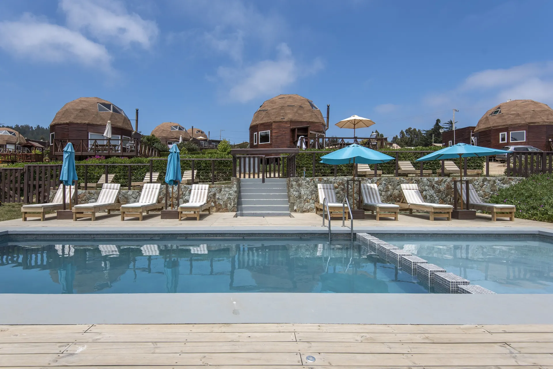 A piscina com espreguiçadeiras e guarda-sóis turquesa, e os domos de madeira atrás.