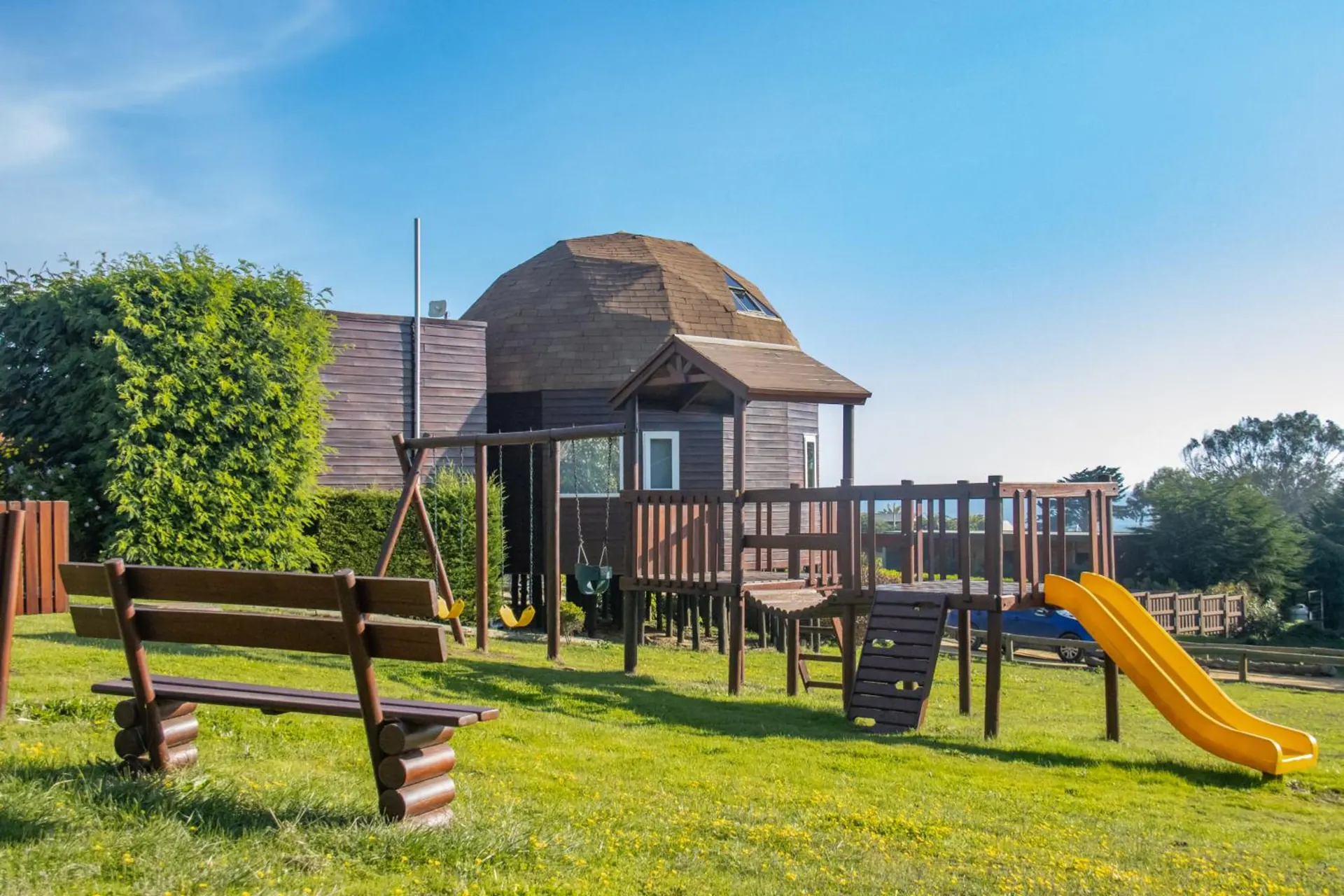 Wooden children's playground with a yellow slide, in front of a dome.