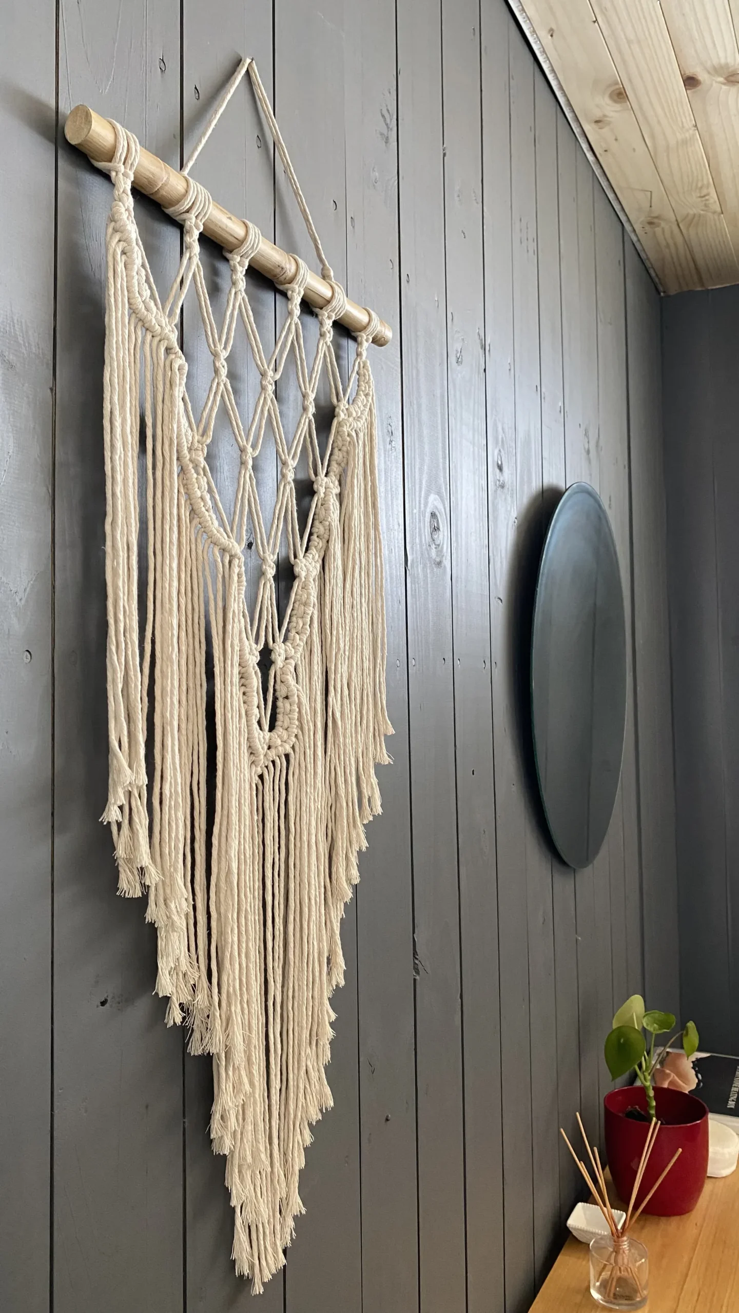 Tapeçaria de macramê e um espelho redondo na parede de madeira cinza da sala de massagem.