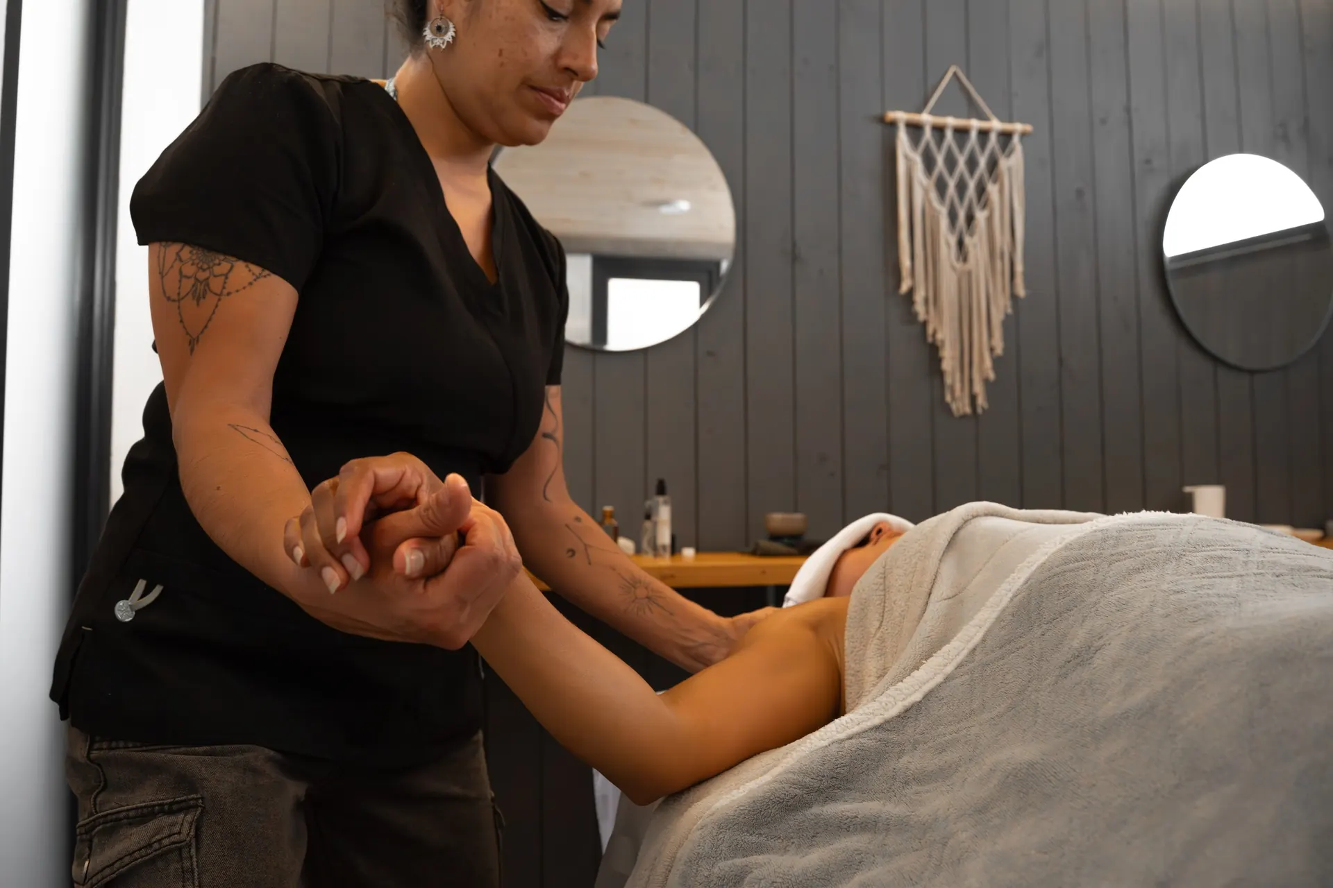 The massage therapist holds the arm of a client lying on the table, with the macramé hanging and round mirrors behind.