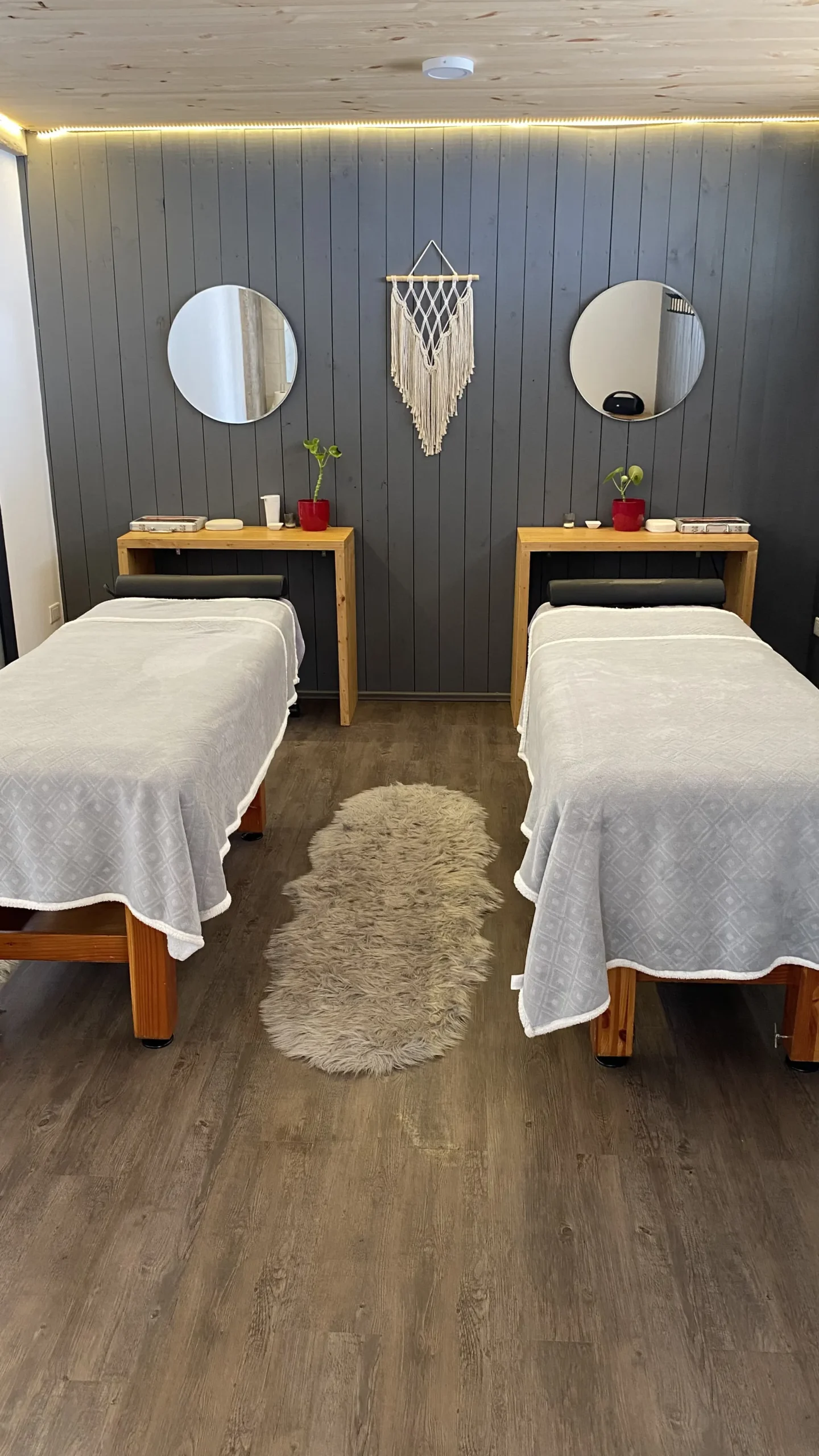Sala de massagem com duas macas, dois espelhos redondos e uma tapeçaria de macramê na parede de madeira cinza.