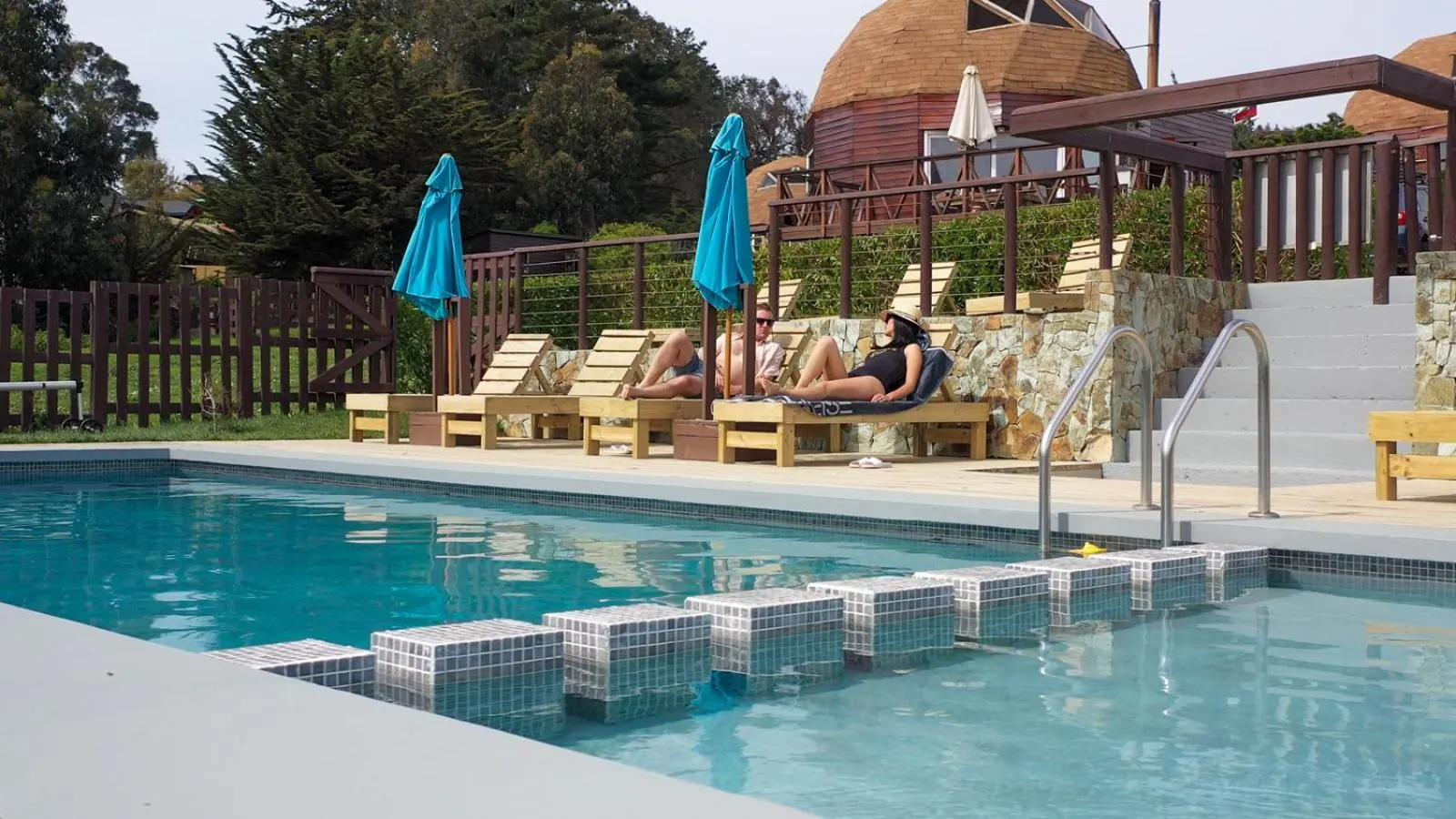 A piscina com um banco de azulejos dentro da água e espreguiçadeiras de madeira no terraço.