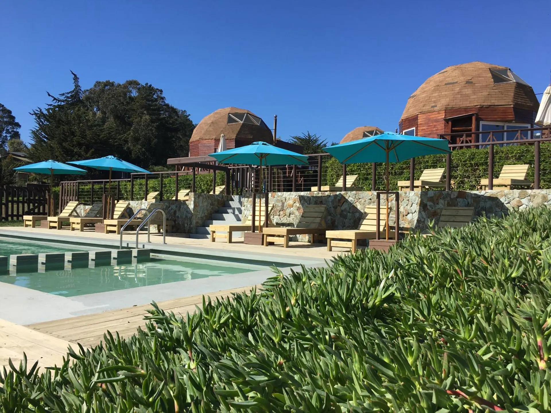 A piscina com espreguiçadeiras e guarda-sóis turquesa, e domos de madeira ao fundo.