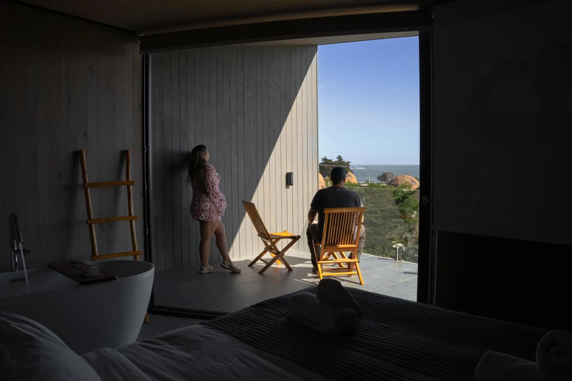 Two people by the large window of a suite look at the sea from the half-light.