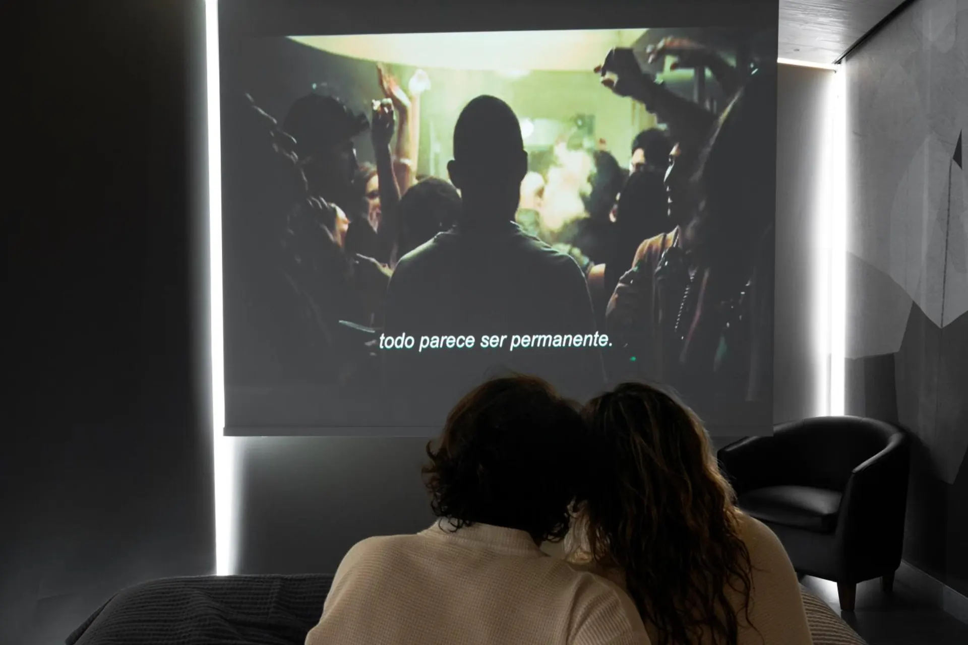 A couple watches a movie projected on the wall of the suite, in the dark.
