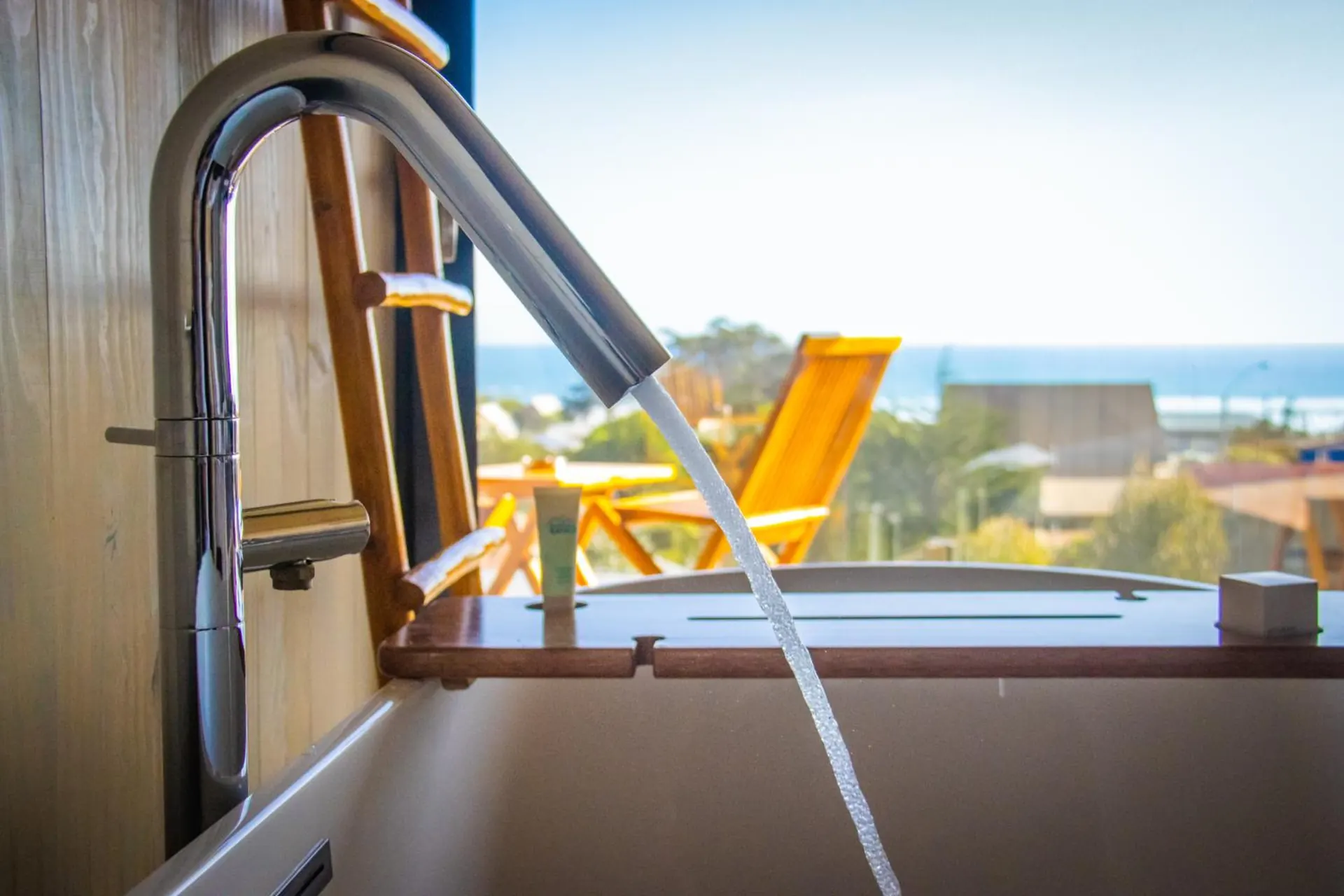 A tub with the faucet stream falling and, through the window, the sea and a yellow chair.