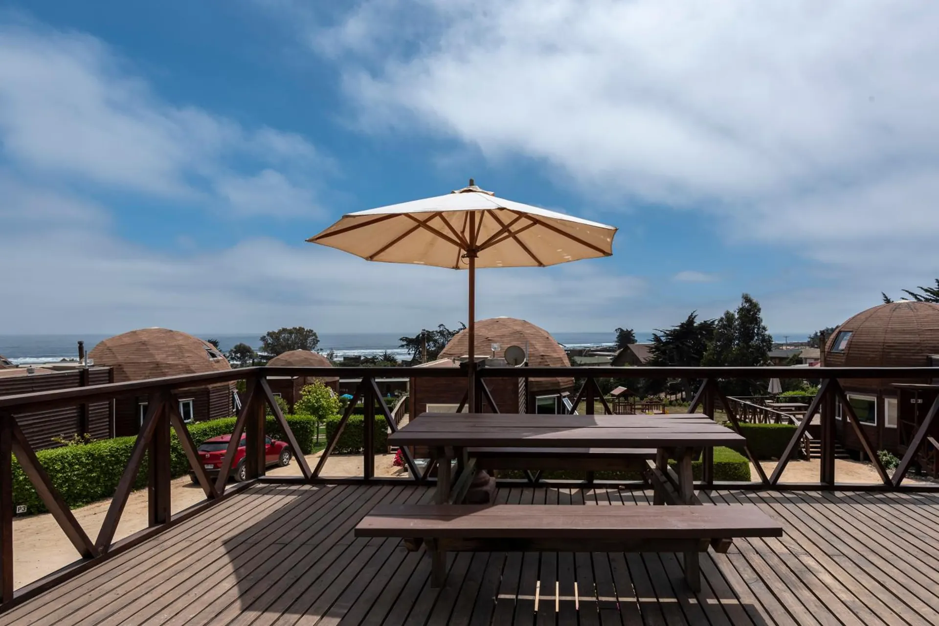 Terraço de madeira com guarda-corpo, uma mesa com guarda-sol e os domos e o mar atrás.