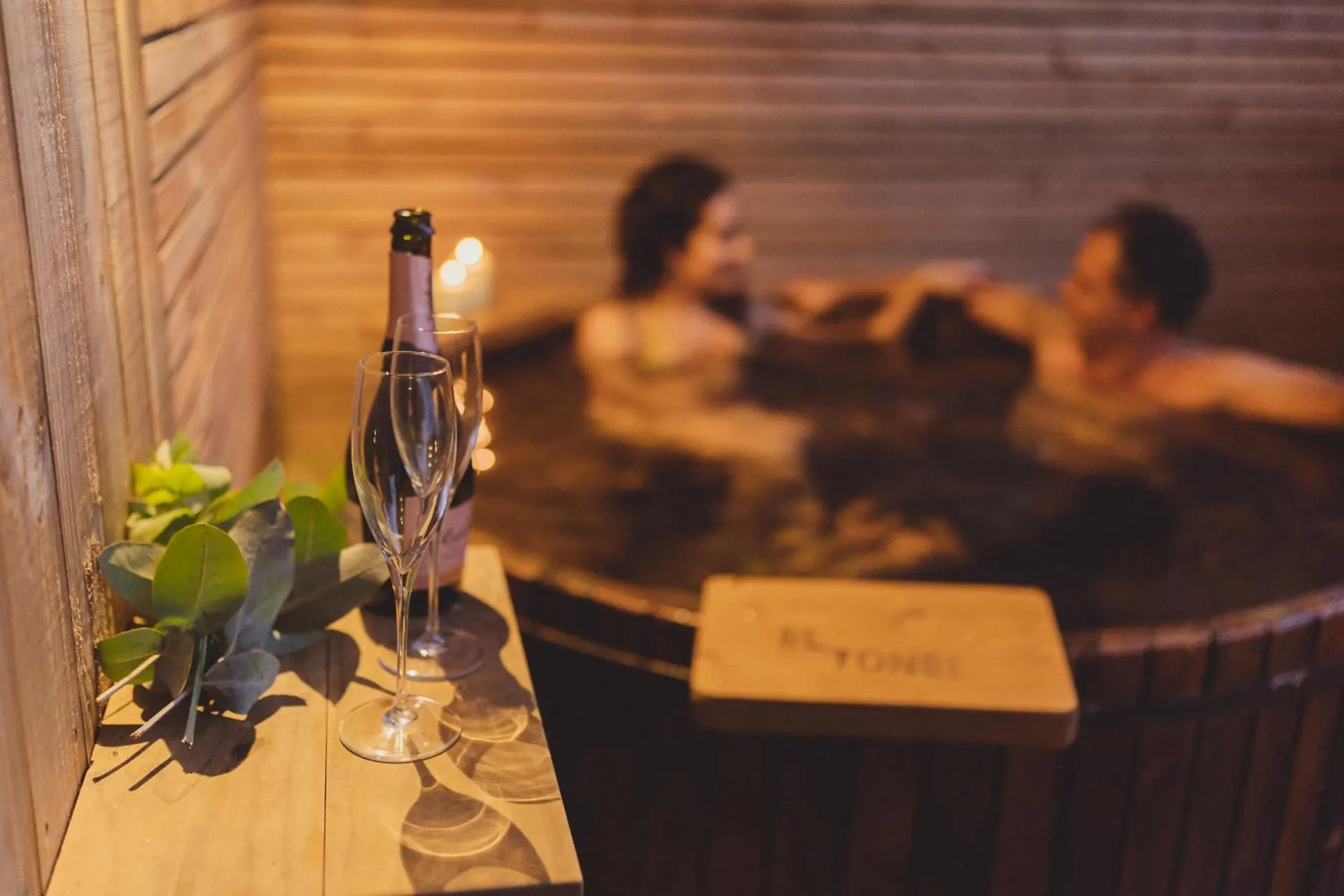 A couple in a wooden hot tub with hot water and glasses of bubbly wine on a board.