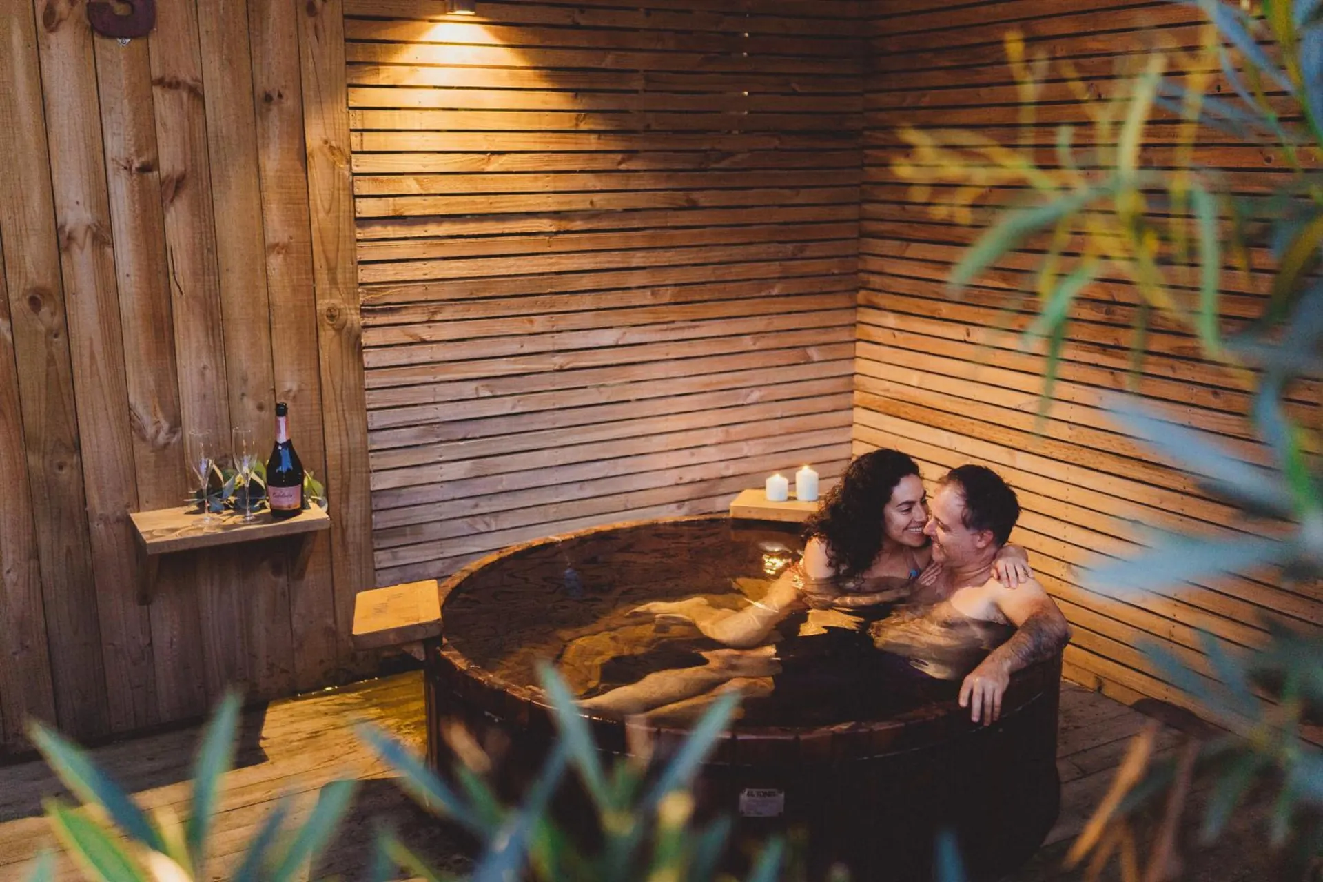 Um casal abraçado dentro de um ofurô de madeira, entre paredes de tábuas.